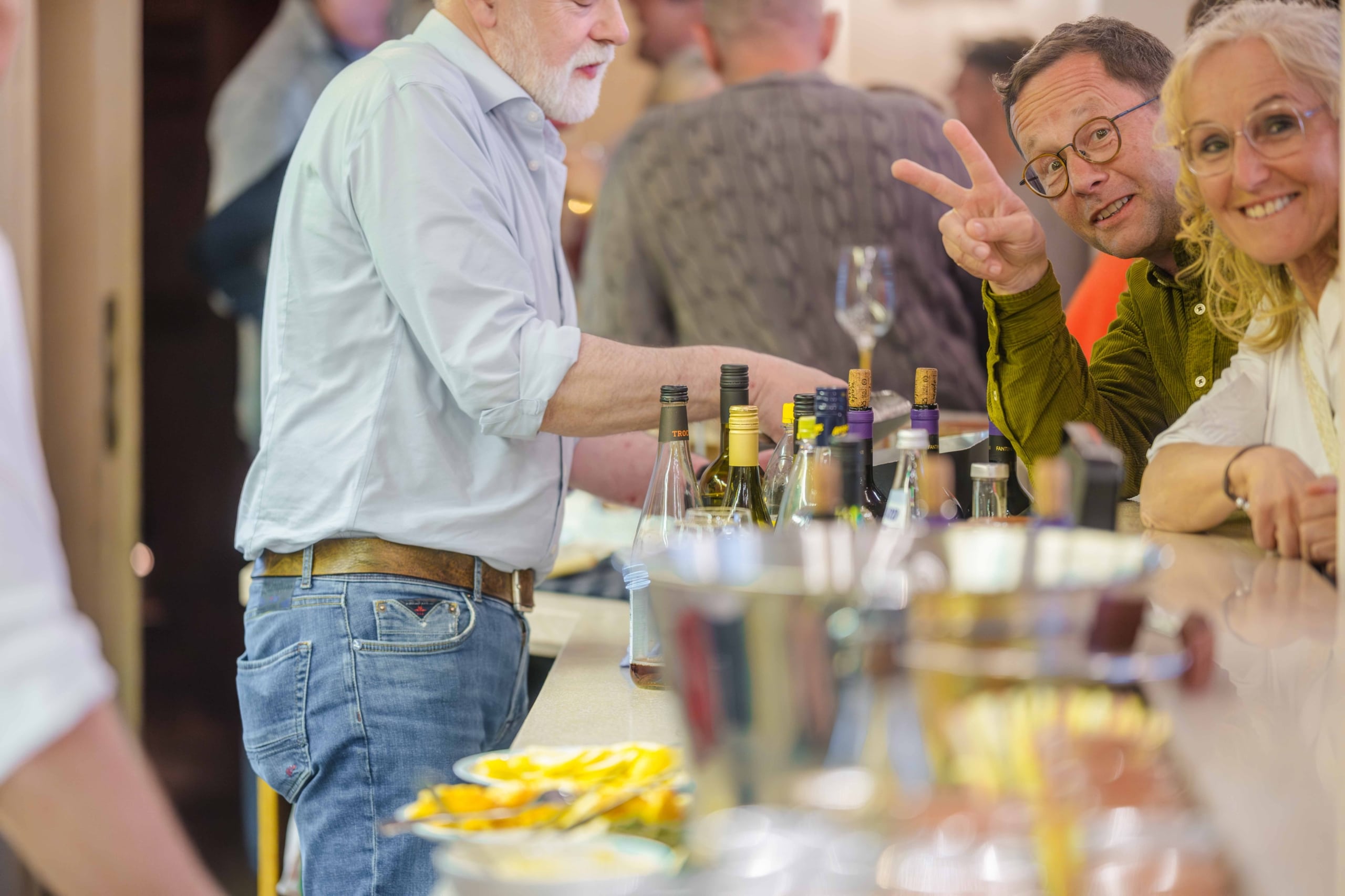 Eine Gruppe von Menschen steht an einer Bar, lächelt und unterhält sich. Ein Mann zeigt das Peace-Zeichen, während auf der Theke Flaschen und Gläser zu sehen sind. Die Atmosphäre wirkt lebhaft und freundlich.